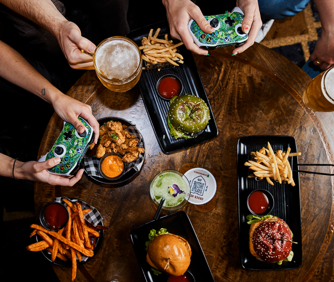 Person playing games using an Xbox controller with fries, a burger, fried Chicken, beers and cocktails on the table
