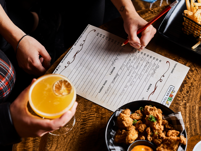 Person filling out a Tavern Trivia answer sheet with fried chicken, a glass of wine and a burger on the table
